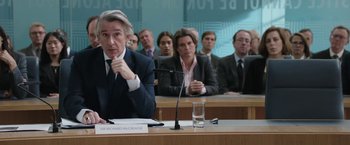 Movie still from “Greed” (2019), directed by Michael Winterbottom – A group of people sitting at a table in front of a microphone; Medium shot, Over the shoulder angle