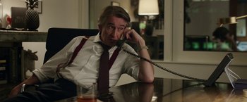 Movie still from “Greed” (2019), directed by Michael Winterbottom – A man sitting at a table talking on a phone; Medium shot, Over the shoulder angle
