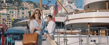 Movie still from “Greed” (2019), directed by Michael Winterbottom – A woman with a purse standing next to a man on a boat; Wide shot, Low angle