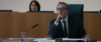 Movie still from “Greed” (2019), directed by Michael Winterbottom – A man sitting in front of a microphone in front of a woman; Medium shot, Over the shoulder angle