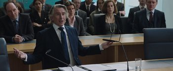 Movie still from “Greed” (2019), directed by Michael Winterbottom – A man in a business suit is giving a presentation; Medium shot, Over the shoulder angle