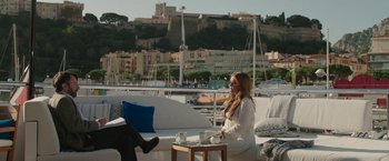 Movie still from “Greed” (2019), directed by Michael Winterbottom – A woman sitting on top of a couch next to a coffee table; Wide shot, Over the shoulder angle