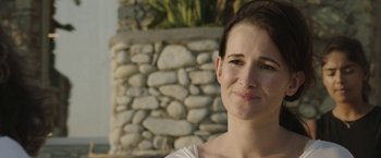 Movie still from “Greed” (2019), directed by Michael Winterbottom – A person smiling near a stone wall; Close Up shot, Over the shoulder angle