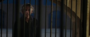 Movie still from “Greed” (2019), directed by Michael Winterbottom – A man standing behind bars in a jail cell; Medium shot, Low angle