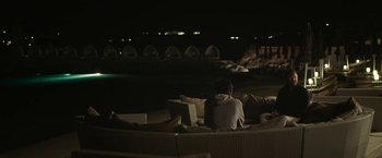 Movie still from “Greed” (2019), directed by Michael Winterbottom – A man sitting on a couch at night with a book; Wide shot, High angle