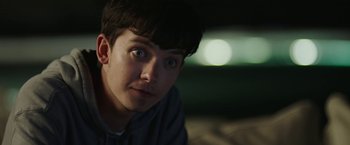 Movie still from “Greed” (2019), directed by Michael Winterbottom – A young man stares into the camera with a blurry background; Close Up shot, Over the shoulder angle