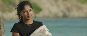 Movie still from “Greed” (2019), directed by Michael Winterbottom – A young woman holding a blanket near a body of water; Close Up shot, Over the shoulder angle