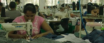 Movie still from “Greed” (2019), directed by Michael Winterbottom – A group of people working in a sewing room; Medium shot, High angle