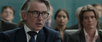 Movie still from “Greed” (2019), directed by Michael Winterbottom – A person wearing a suit and glasses; Close Up shot, Over the shoulder angle