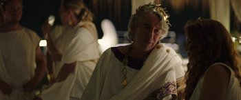 Movie still from “Greed” (2019), directed by Michael Winterbottom – An older man wearing a crown and a white robe; Close Up shot, Over the shoulder angle