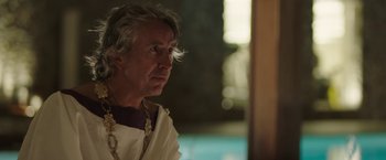 Movie still from “Greed” (2019), directed by Michael Winterbottom – An older man wearing a white shirt and a gold necklace; Close Up shot, Over the shoulder angle