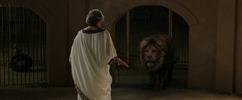 Movie still from “Greed” (2019), directed by Michael Winterbottom – A man in a robe and a lion in an enclosure; Wide shot, Over the shoulder angle