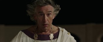 Movie still from “Greed” (2019), directed by Michael Winterbottom – An older man wearing a roman outfit with a beard; Close Up shot, Low angle