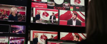 Movie still from “Greed” (2019), directed by Michael Winterbottom – A television screen showing a man and a woman in front of a clock; Medium shot, Over the shoulder angle