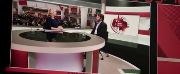 Movie still from “Greed” (2019), directed by Michael Winterbottom – A man and a woman sitting in front of a news desk; Wide shot, High angle