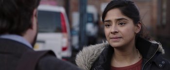 Movie still from “Greed” (2019), directed by Michael Winterbottom – A young woman is sitting on the sidewalk; Close Up shot, Over the shoulder angle