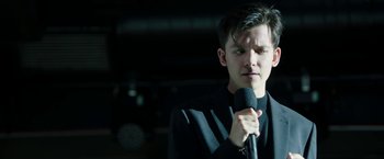 Movie still from “Greed” (2019), directed by Michael Winterbottom – A man holding a microphone in his hand; Close Up shot, Low angle