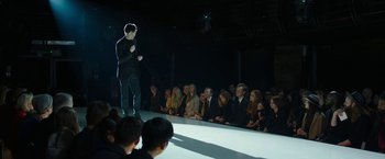 Movie still from “Greed” (2019), directed by Michael Winterbottom – A man standing on a stage in front of a group of people; Wide shot, Over the shoulder angle