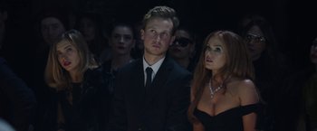 Movie still from “Greed” (2019), directed by Michael Winterbottom – A group of people in a dark room; Medium shot, Low angle