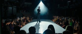 Movie still from “Greed” (2019), directed by Michael Winterbottom – A man standing on a runway with a microphone in his hand; Wide shot, High angle