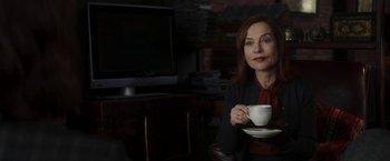 Movie still from “Greta” (2018), directed by Neil Jordan – A woman holding a coffee cup while sitting in front of a television; Medium shot, Over the shoulder angle