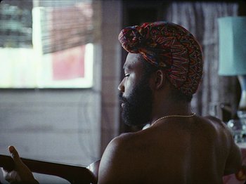 Movie still from “Guava Island” (2019), directed by Hiro Murai – A man with a turban sitting in a chair; Close Up shot, Over the shoulder angle