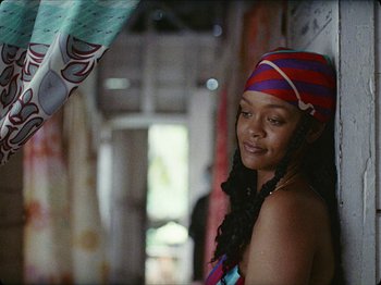 Movie still from “Guava Island” (2019), directed by Hiro Murai – A woman wearing a colorful head scarf and a blue and red striped hat; Close Up shot, Low angle