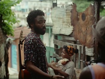 Movie still from “Guava Island” (2019), directed by Hiro Murai – A man standing next to a woman holding a guitar; Medium shot, Over the shoulder angle