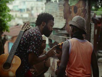 Movie still from “Guava Island” (2019), directed by Hiro Murai – A group of people standing around talking to each other; Medium shot, Over the shoulder angle