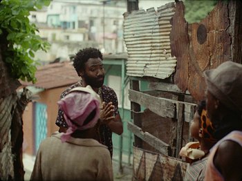 Movie still from “Guava Island” (2019), directed by Hiro Murai – A group of people standing next to each other on a street; Medium shot, Over the shoulder angle