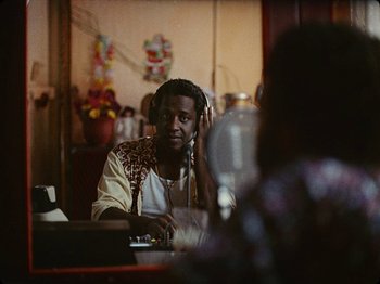 Movie still from “Guava Island” (2019), directed by Hiro Murai – A man sitting at a table with a microphone in front of him; Medium shot, Over the shoulder angle