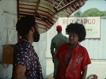 Movie still from “Guava Island” (2019), directed by Hiro Murai – A man and a woman standing under an umbrella; Medium shot, Over the shoulder angle