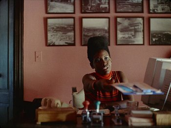 Movie still from “Guava Island” (2019), directed by Hiro Murai – A woman sitting at a table in front of a computer; Medium shot, Over the shoulder angle