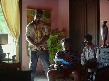 Movie still from “Guava Island” (2019), directed by Hiro Murai – Three people are sitting in a living room; Medium shot, Over the shoulder angle