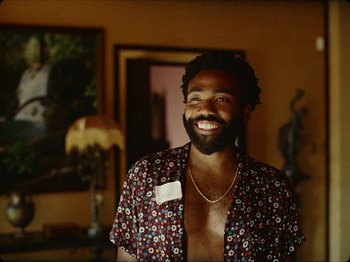 Movie still from “Guava Island” (2019), directed by Hiro Murai – A man smiling for the camera while wearing a floral shirt; Medium shot, Over the shoulder angle