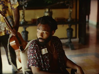 Movie still from “Guava Island” (2019), directed by Hiro Murai – A man sitting in a chair in a room with a guitar; Medium shot, Over the shoulder angle