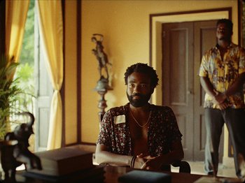 Movie still from “Guava Island” (2019), directed by Hiro Murai – A man sitting at a table in front of a door; Medium shot, Over the shoulder angle