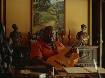 Movie still from “Guava Island” (2019), directed by Hiro Murai – A man sitting at a table holding a guitar; Medium shot, Low angle