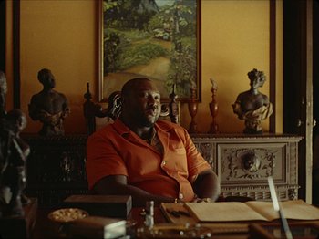 Movie still from “Guava Island” (2019), directed by Hiro Murai – A man sitting at a table in a room; Medium shot, Low angle