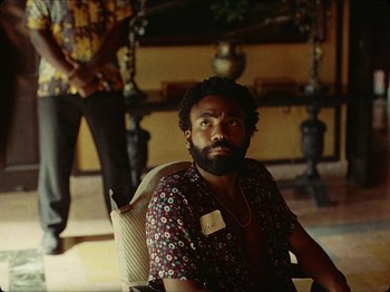 Movie still from “Guava Island” (2019), directed by Hiro Murai – A man sitting in a chair in front of a mirror; Medium shot, Over the shoulder angle
