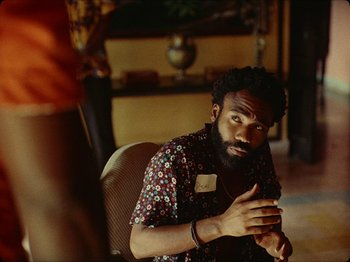 Movie still from “Guava Island” (2019), directed by Hiro Murai – A man sitting in front of a camera in a living room; Medium shot, Over the shoulder angle