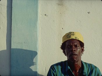 Movie still from “Guava Island” (2019), directed by Hiro Murai – A man wearing a yellow hat standing in front of a wall; Close Up shot, Low angle