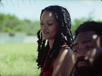 Movie still from “Guava Island” (2019), directed by Hiro Murai – A woman with long black hair is standing next to a man with long black hair; Close Up shot, Over the shoulder angle