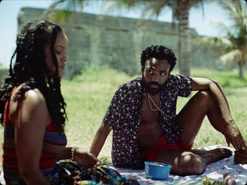Movie still from “Guava Island” (2019), directed by Hiro Murai – A man and a woman sitting on a blanket in the grass; Medium shot, Over the shoulder angle