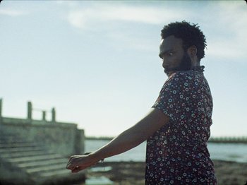 Movie still from “Guava Island” (2019), directed by Hiro Murai – A man standing in front of a body of water; Medium shot, Over the shoulder angle