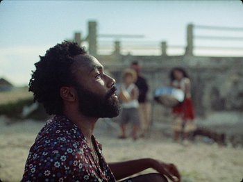 Movie still from “Guava Island” (2019), directed by Hiro Murai – A man sitting on the ground with his eyes wide open; Medium shot, Over the shoulder angle