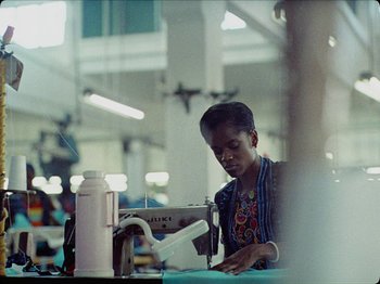 Movie still from “Guava Island” (2019), directed by Hiro Murai – A person sitting at a sewing machine in a room; Medium shot, Low angle