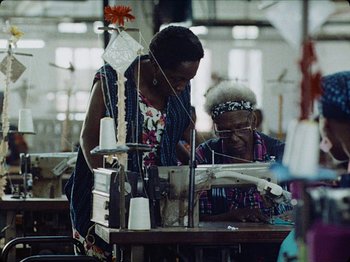 Movie still from “Guava Island” (2019), directed by Hiro Murai – Two women working in an industrial sewing machine; Medium shot, Over the shoulder angle
