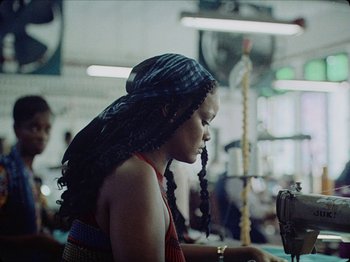 Movie still from “Guava Island” (2019), directed by Hiro Murai – A woman with a scarf on her head is sitting in a sewing room; Medium shot, Low angle