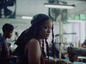 Movie still from “Guava Island” (2019), directed by Hiro Murai – A young woman sitting at a sewing machine in a room; Medium shot, Low angle